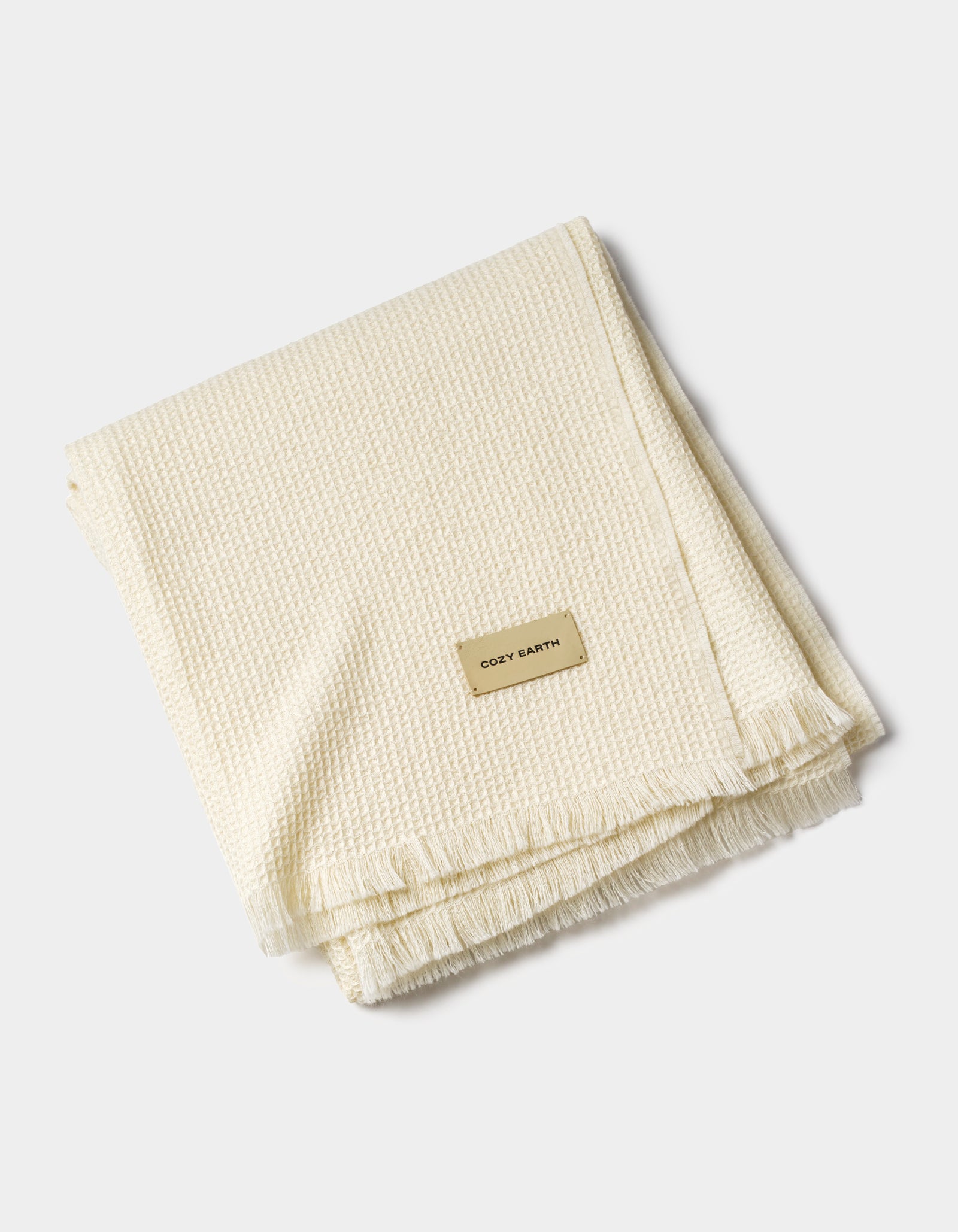 A folded, cream-colored Alpaca Waffle Throw with a textured design, fringe edges, and a rectangular "Cozy Earth" label, displayed on a plain white background.