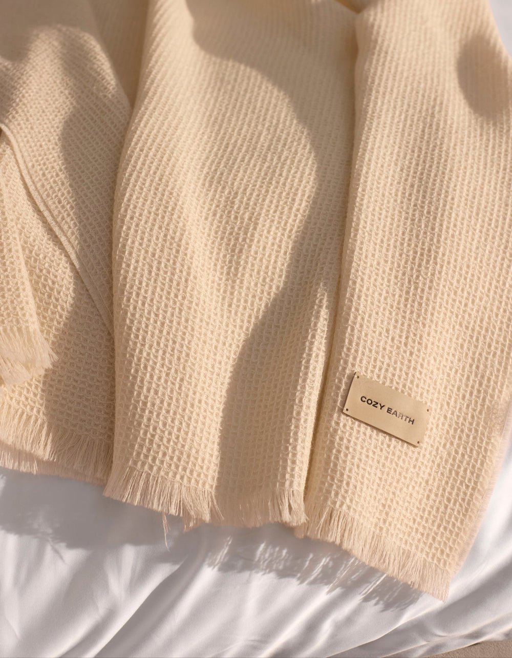 A Cozy Earth Alpaca Waffle Throw in creme, featuring a textured waffle weave and fringed edges, is draped over a white surface. A beige tag on the throw displays the brand name "Cozy Earth.