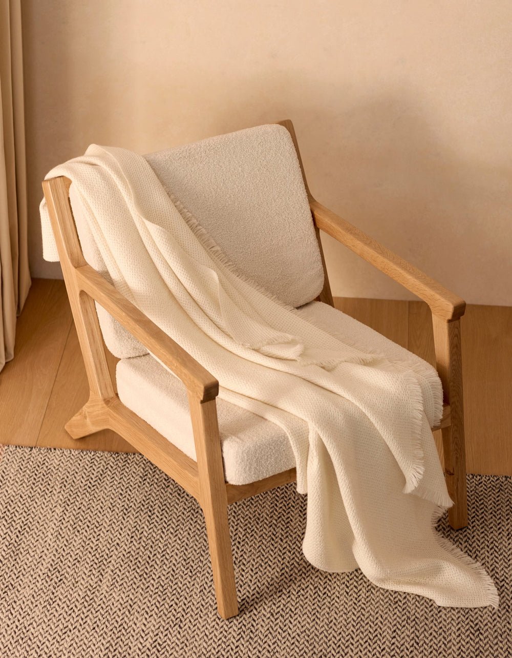 A modern wooden armchair with light beige upholstery sits on a herringbone rug, draped with the Cozy Earth Alpaca Waffle Throw. The soft blanket and minimal setting create a warm, inviting atmosphere.