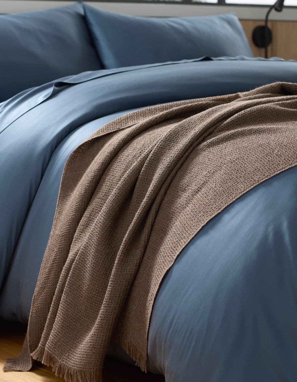 A close-up of a neatly made bed with smooth blue bedding and a Cozy Earth Alpaca Waffle Throw in brown draped casually over the edge.