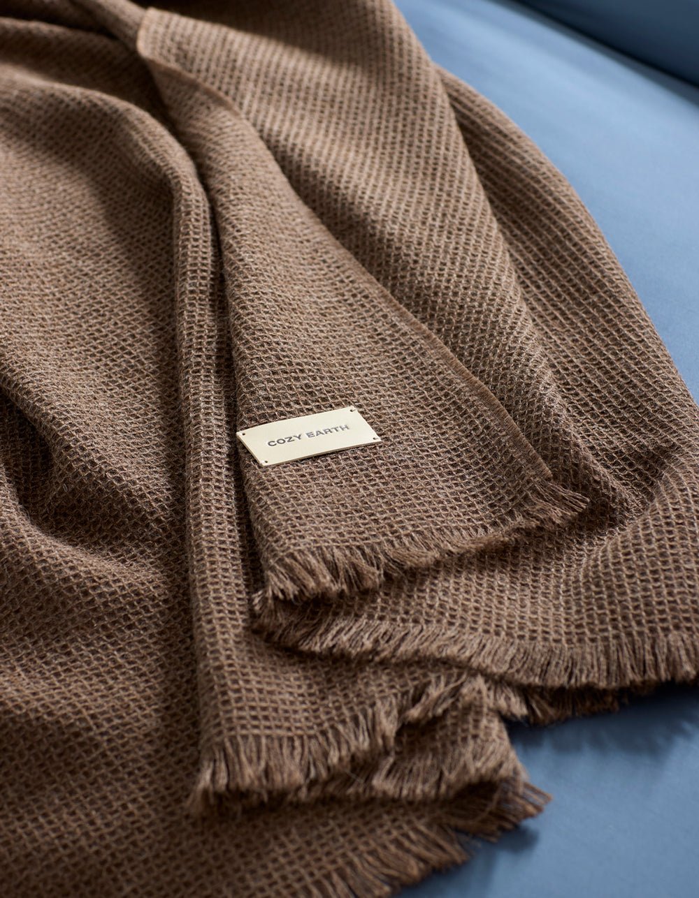A brown Alpaca Waffle Throw by Cozy Earth, featuring a textured weave and fringed edges, is draped over a blue surface. A small white label with the brand name "Cozy Earth" is visible on the blanket.