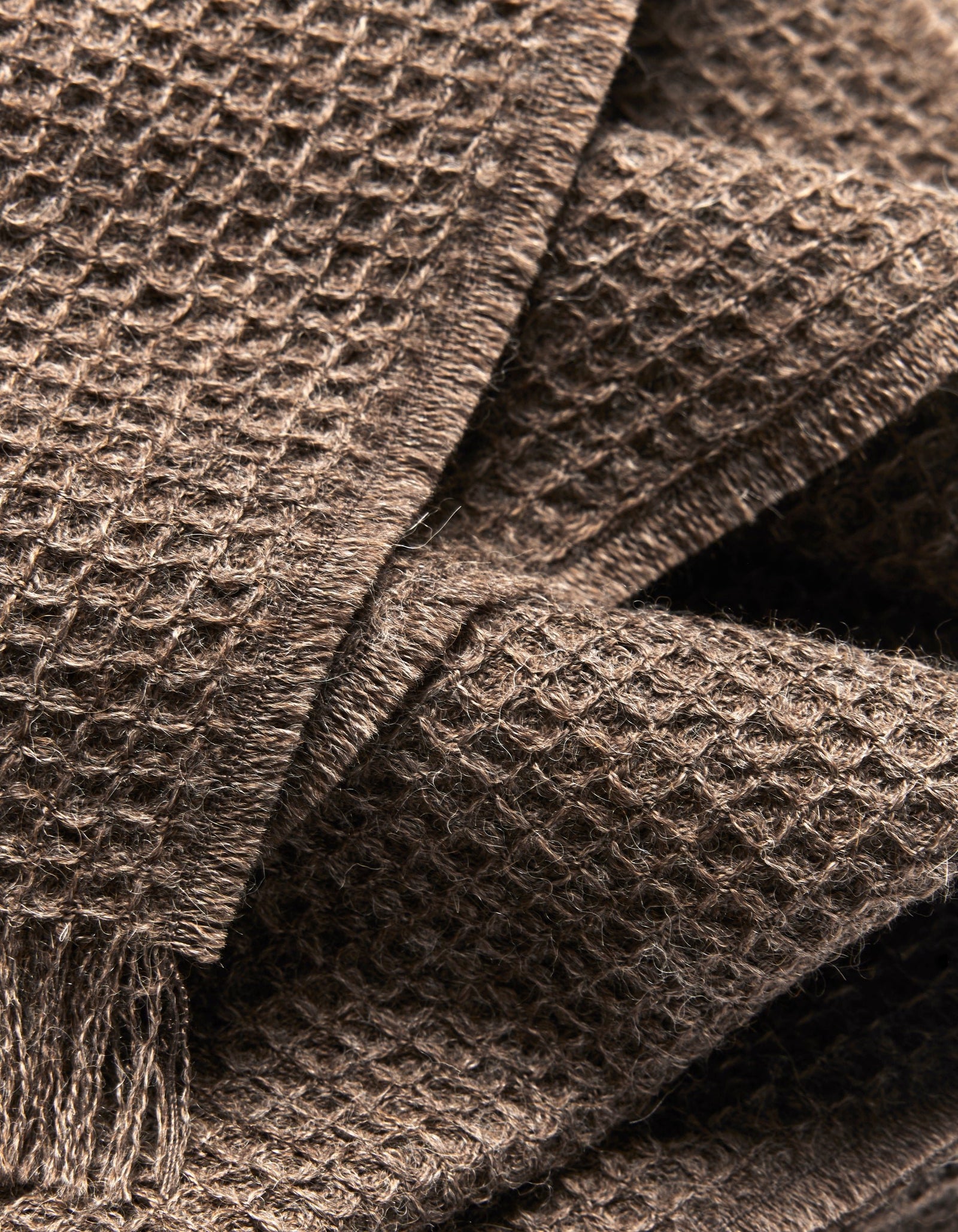 Close-up of the Alpaca Waffle Throw by Cozy Earth, highlighting its brown waffle weave texture, soft fibers, and layered folds. Fringed edges are visible in the lower left corner, emphasizing its cozy appeal.