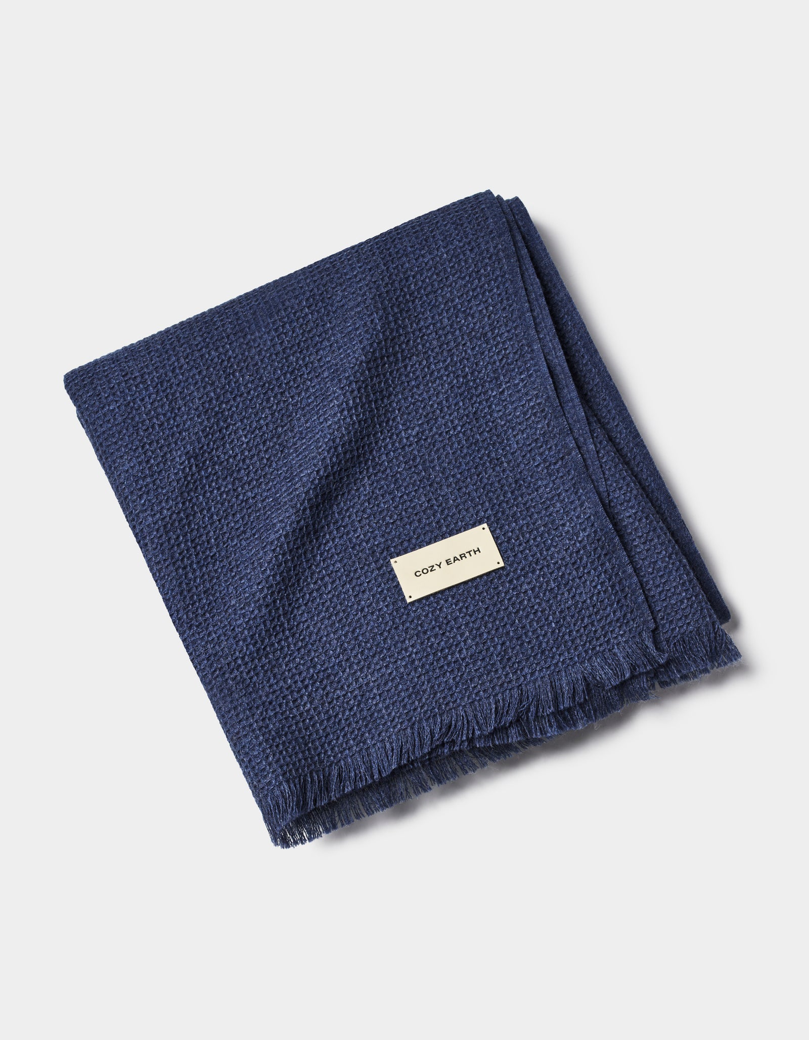 A folded Alpaca Waffle Throw from Cozy Earth in dark blue, featuring a textured pattern, fringed edges, and a small rectangular "Cozy Earth" label sewn near one corner.