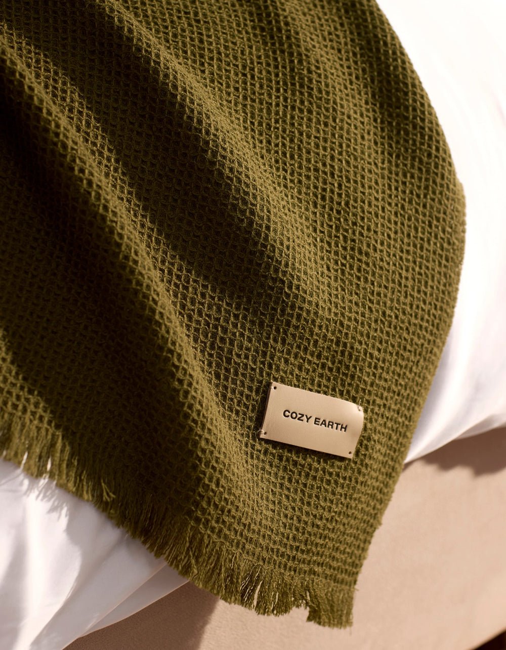 A Cozy Earth Alpaca Waffle Throw in dark green, featuring a textured waffle weave and fringed edges, is draped over a white surface. A beige tag on the throw displays the brand name "Cozy Earth.