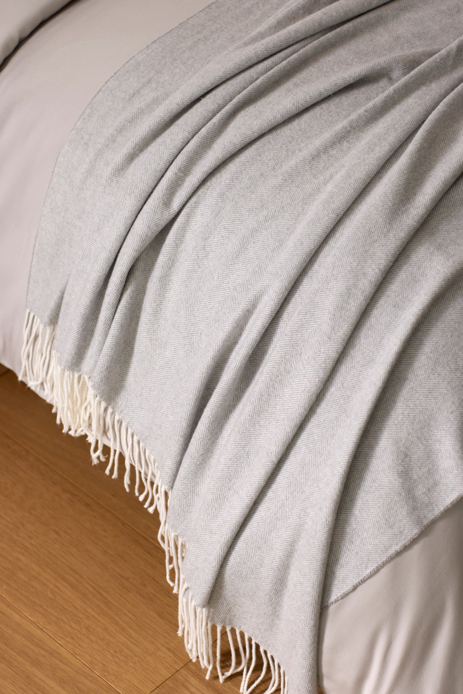 The Cozy Earth Bamboo Herringbone Tassel Throw in light gray is draped neatly over the edge of a bed with white bedding, set on a wooden floor.