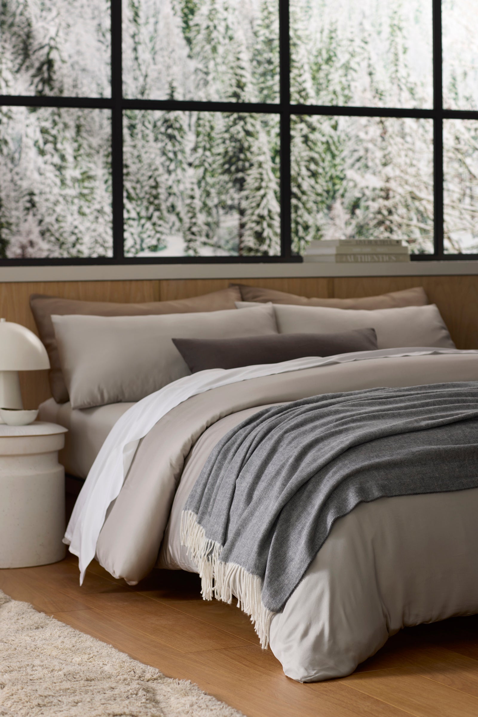 A cozy bed with gray and beige bedding sits by a large window overlooking a snowy forest. A Bamboo Herringbone Tassel Throw by Cozy Earth is draped over the bed, and a white lamp rests on the bedside table, creating a warm, inviting atmosphere.