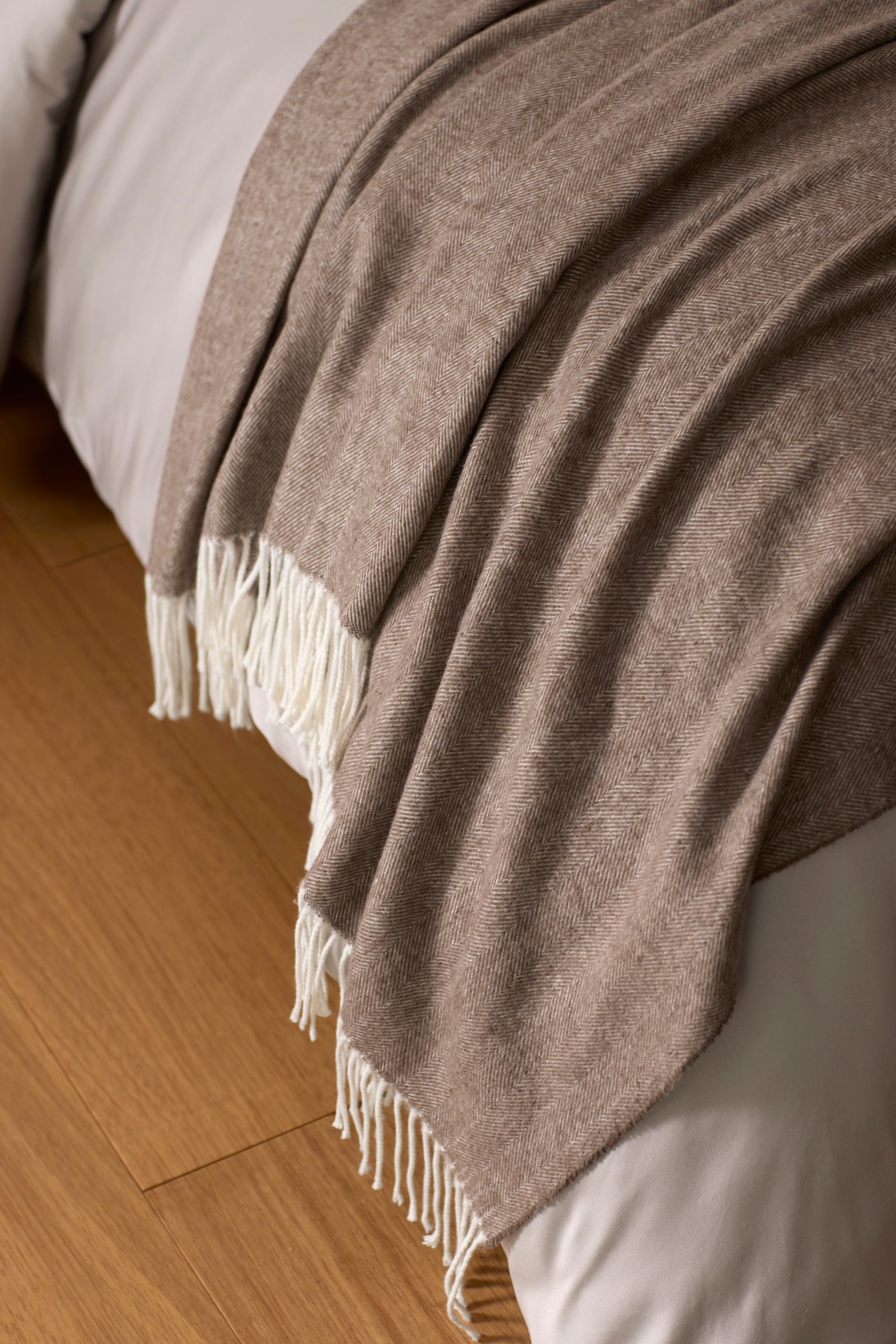 The Cozy Earth Bamboo Herringbone Tassel Throw, a soft light brown blanket with white fringe, is draped over a white bed resting on a wooden floor.