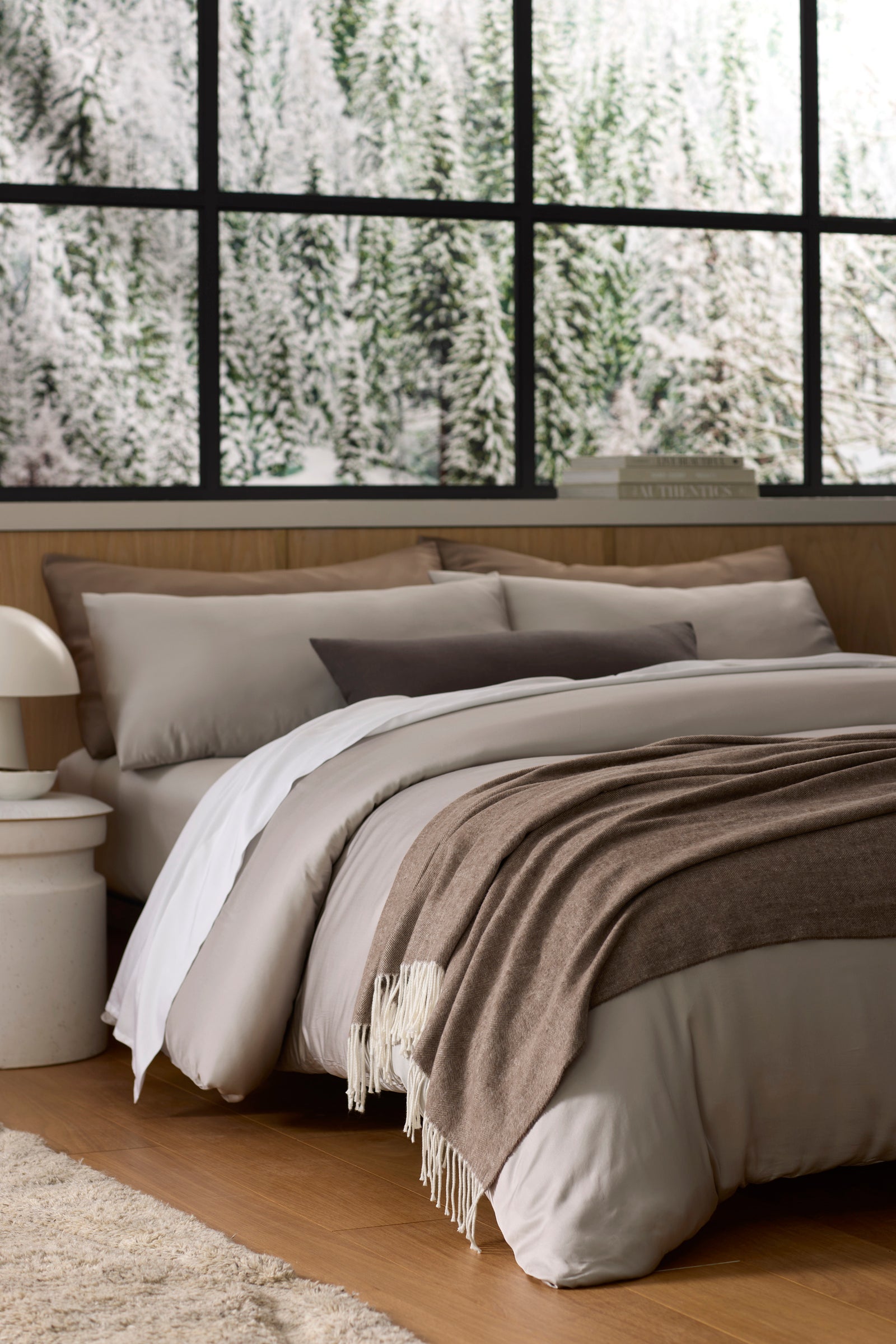 A cozy bed features Cozy Earth's Bamboo Herringbone Tassel Throw in beige and brown, set in a modern room with large windows framing a snowy forest view.