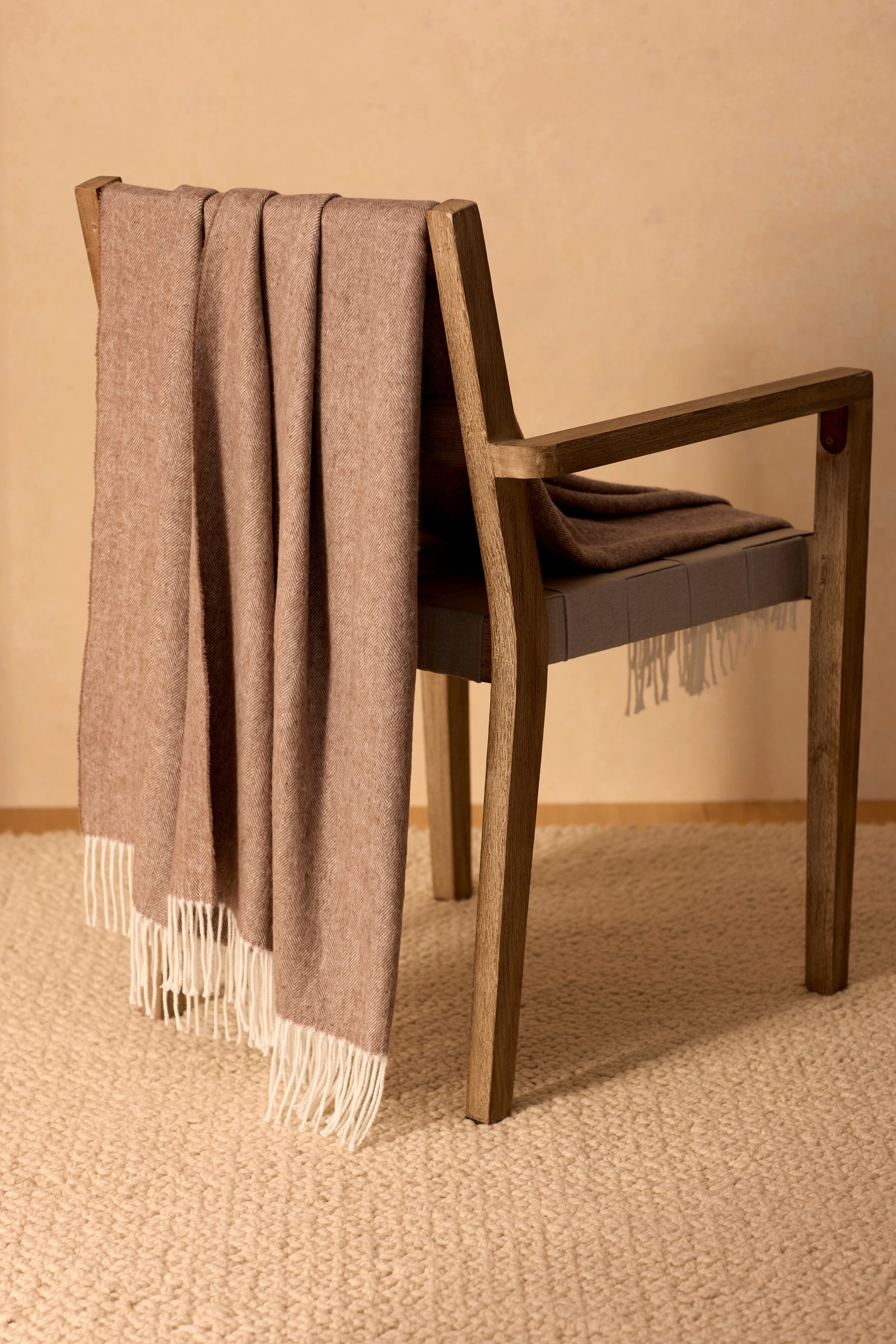 A wooden chair with the Cozy Earth Bamboo Herringbone Tassel Throw in taupe, featuring white fringed ends, is draped over the backrest and sits on a textured beige carpet against a light tan wall.
