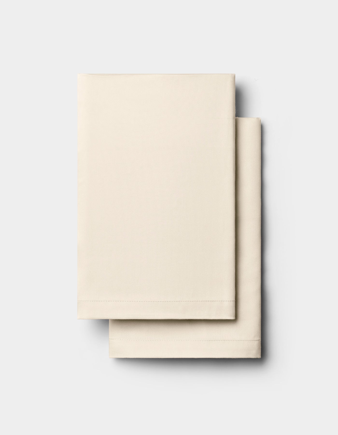 Two neatly folded, cream-colored rectangular Bamboo Pillowcases by Cozy Earth are stacked on top of each other against a plain, light gray background.|Color:Creme