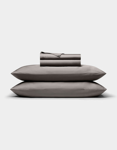The Cozy Earth Bamboo Sheet Set, in dark gray, is neatly folded with matching pillowcases stacked atop two pillows, all arranged against a plain white background.|Color:Charcoal