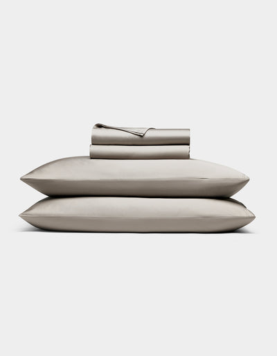 The Cozy Earth Bamboo Sheet Set, in light gray, includes neatly folded bed sheets and pillowcases stacked atop two pillows, all shown against a plain white background.|Color:Dove Grey