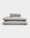 The Bamboo Sheet Set by Cozy Earth in light gray is neatly folded, with two pillows stacked and both flat and fitted sheets rolled on top, all displayed against a white background.|Color:Light Grey