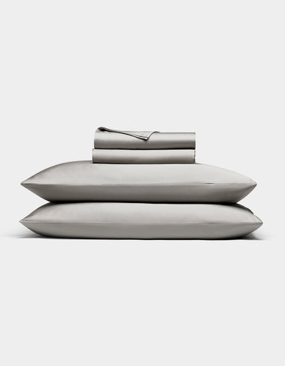 The Bamboo Sheet Set by Cozy Earth in light gray is neatly folded, with two pillows stacked and both flat and fitted sheets rolled on top, all displayed against a white background.|Color:Light Grey