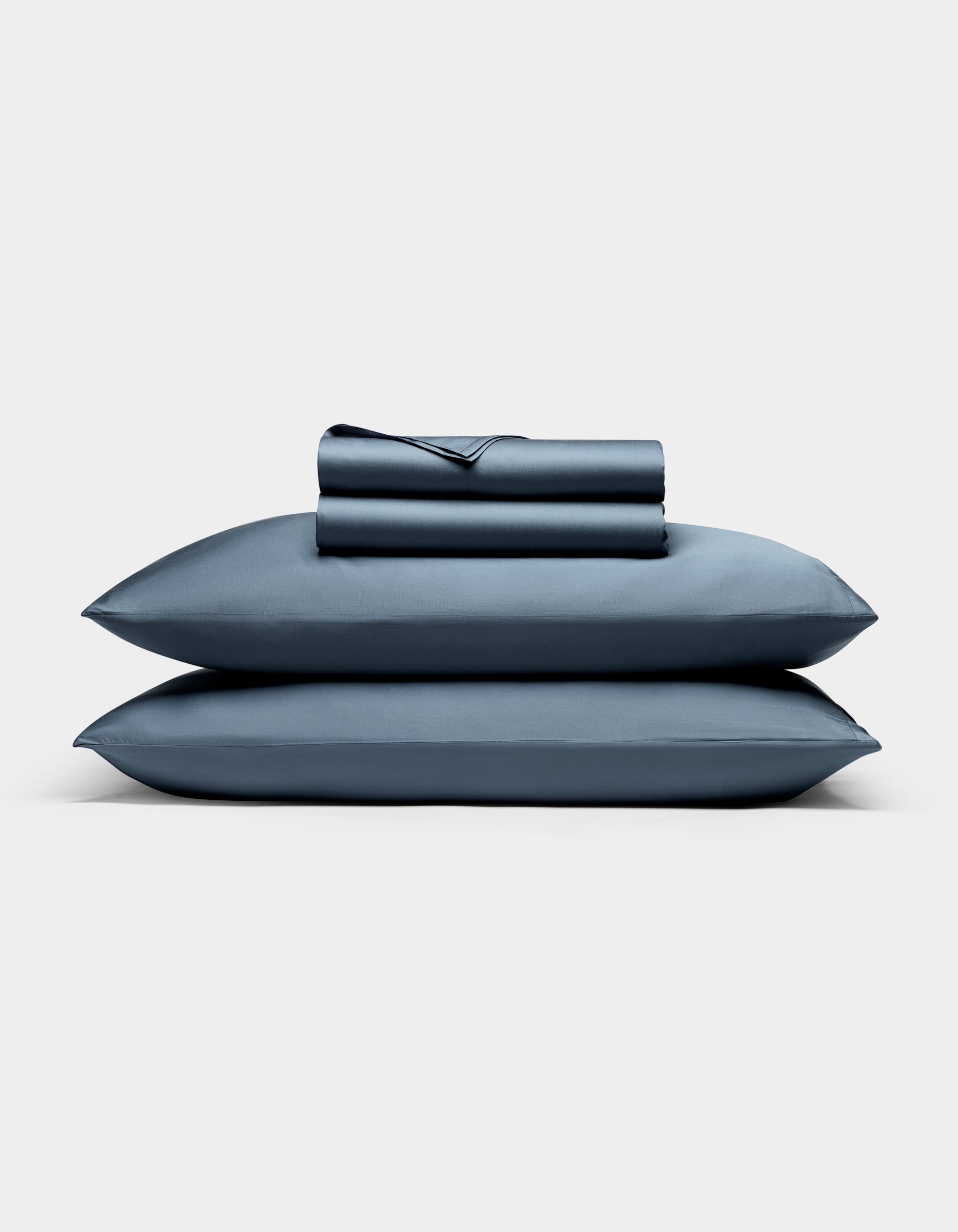 A neatly folded Cozy Earth Bamboo Sheet Set, featuring a fitted sheet, flat sheet, and two pillowcases, is stacked atop two matching blue pillows against a plain white background.|Color:Pacific Blue