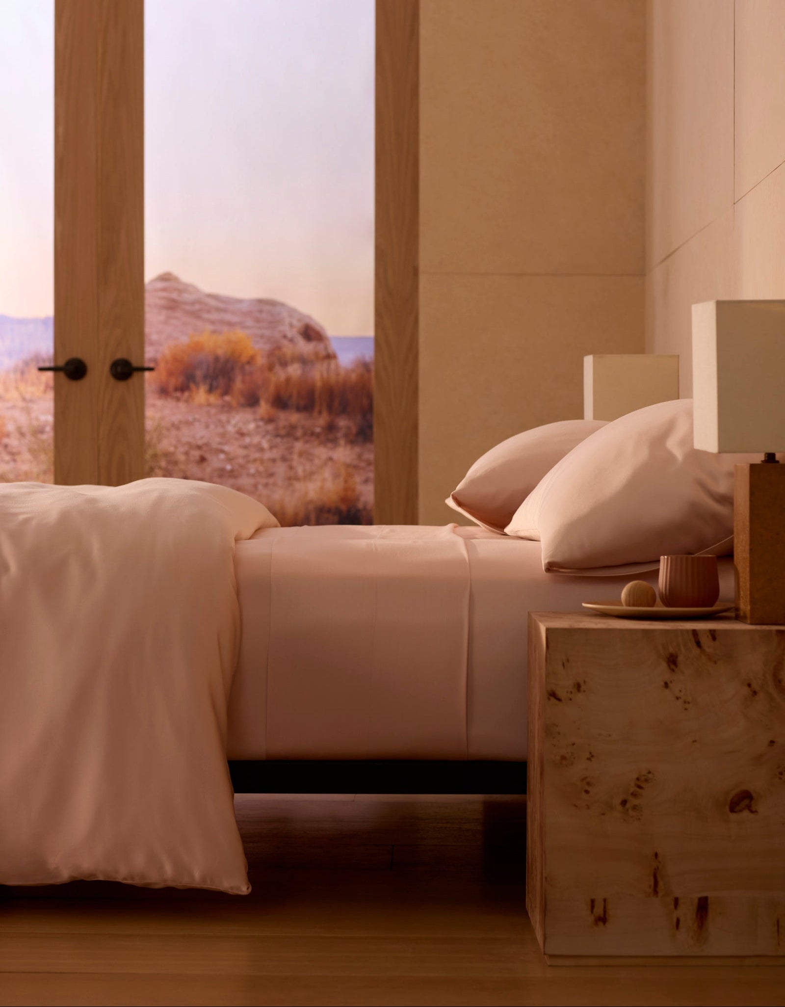 A neatly made bed with Cozy Earth's Bamboo Sheet Set in light pink sits next to a wooden nightstand with a lamp. Large windows showcase a scenic sunlit desert, adding to the minimalist, cozy atmosphere.