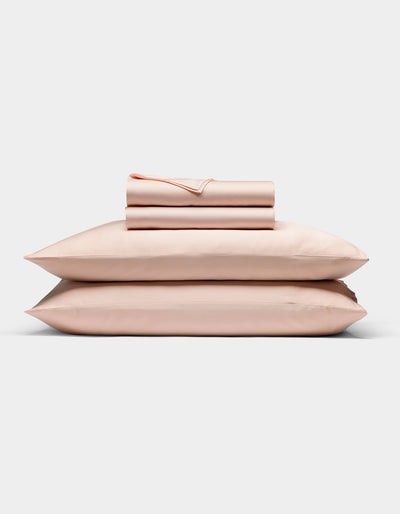 The Cozy Earth Bamboo Sheet Set in light pink is neatly folded with matching pillowcases, stacked on two pillows against a plain white background.|Color:Peony