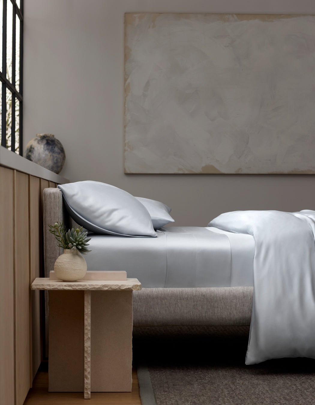 A neatly made bed features Cozy Earth's Bamboo Fitted Sheet in light gray, with matching pillows. A small wooden nightstand holds a round vase with a succulent. Large abstract art hangs above, and natural light streams from the left window.