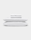 A neatly folded Bamboo Sheet Set by Cozy Earth is stacked on two white pillows, all set against a plain light gray background.|Color:White