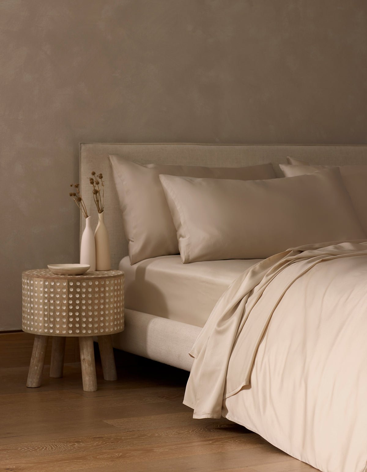 A minimalist bedroom features a beige upholstered bed dressed in Cozy Earth’s Bamboo Flat Sheet, accented with beige pillows. A round wooden nightstand holds two simple vases with dried flowers, complementing the warm, neutral color palette.