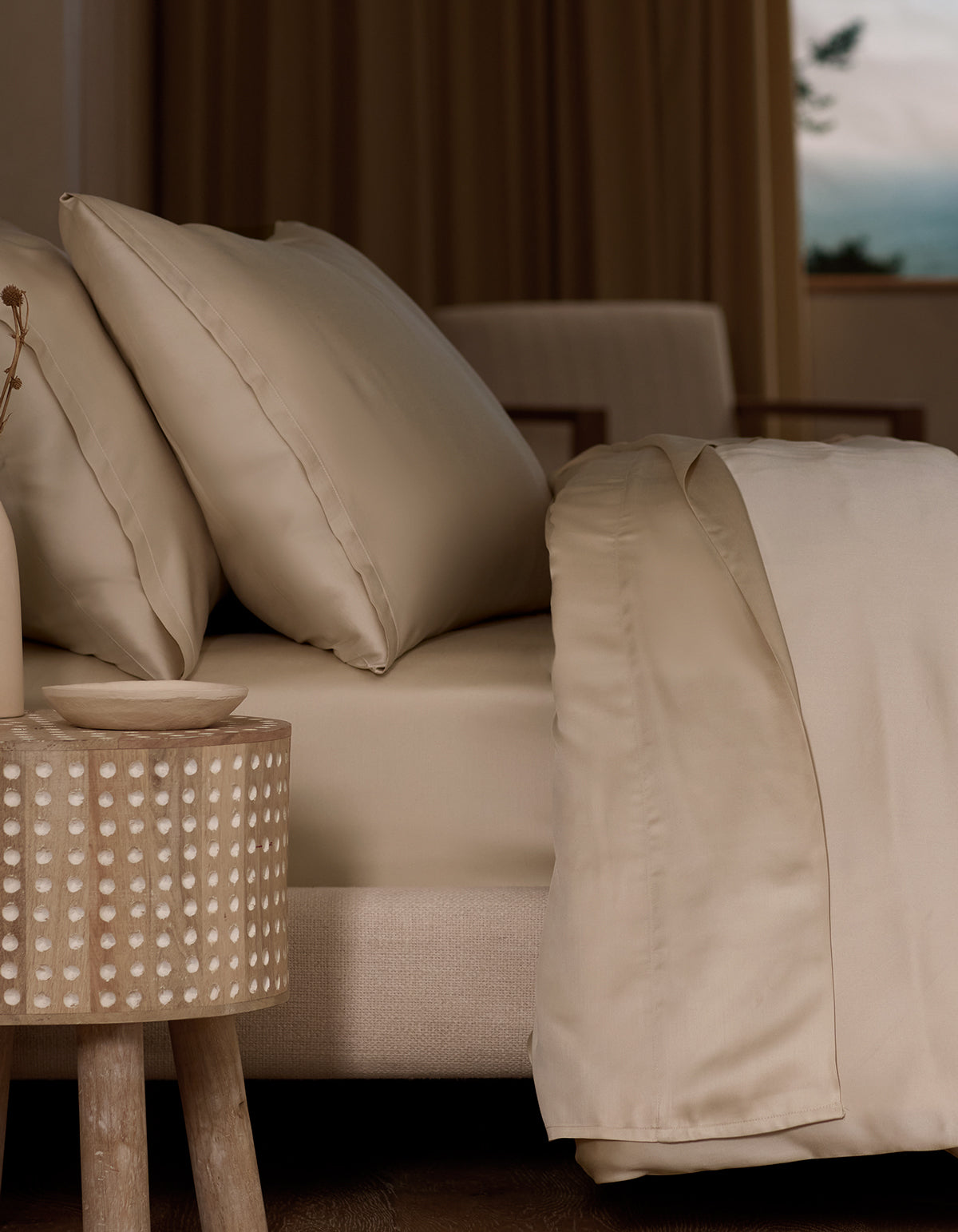 A neatly made bed features Cozy Earth Bamboo Pillowcases in beige, next to a round wooden bedside table with a dotted pattern in a softly lit, neutral room. A window in the background reveals an outdoor view.