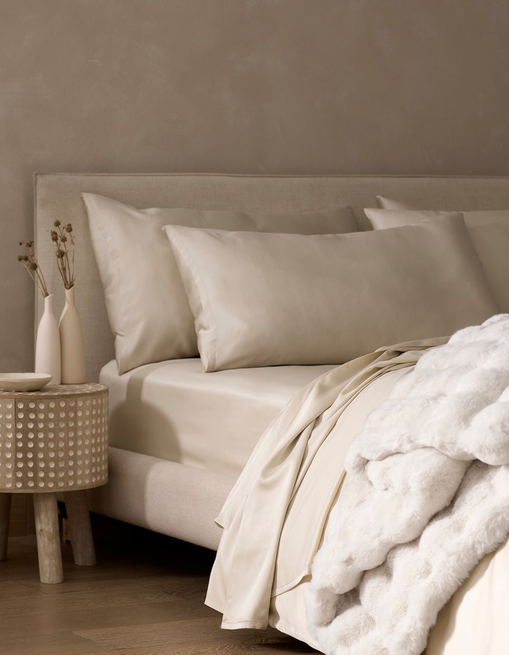 A neatly made bed features Cozy Earth Bamboo Pillowcases in cream, a soft white blanket, and a nightstand with minimal vases of dried plants against a beige wall.|Color:Oat