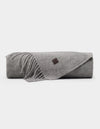 The Cozy Earth Cashmere Tassel Throw, a light gray wool blanket with fringed edges and a small brown leather patch, is neatly rolled and displayed on a plain white background.|Color:Pebble