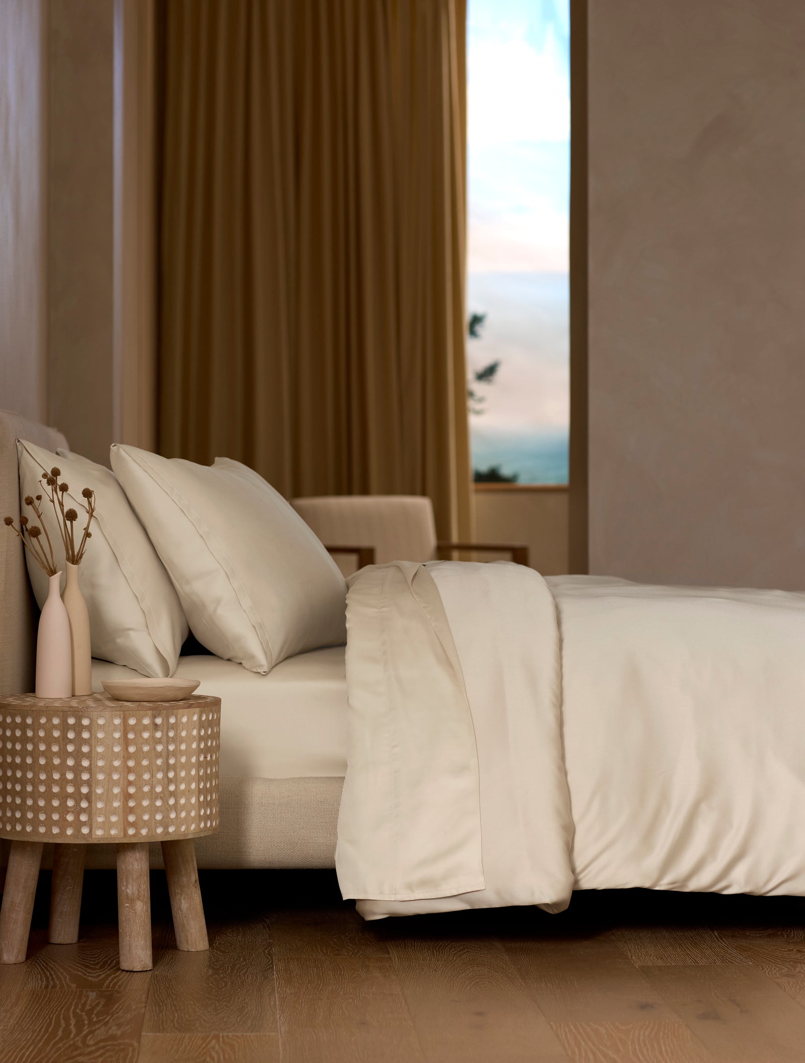 A bed dressed in Cozy Earth’s Bamboo Bedding Deluxe Bundle features cream-colored sheets and pillows, set beside a round wooden table with dried flowers. In the background are a beige armchair and a window with gold curtains.