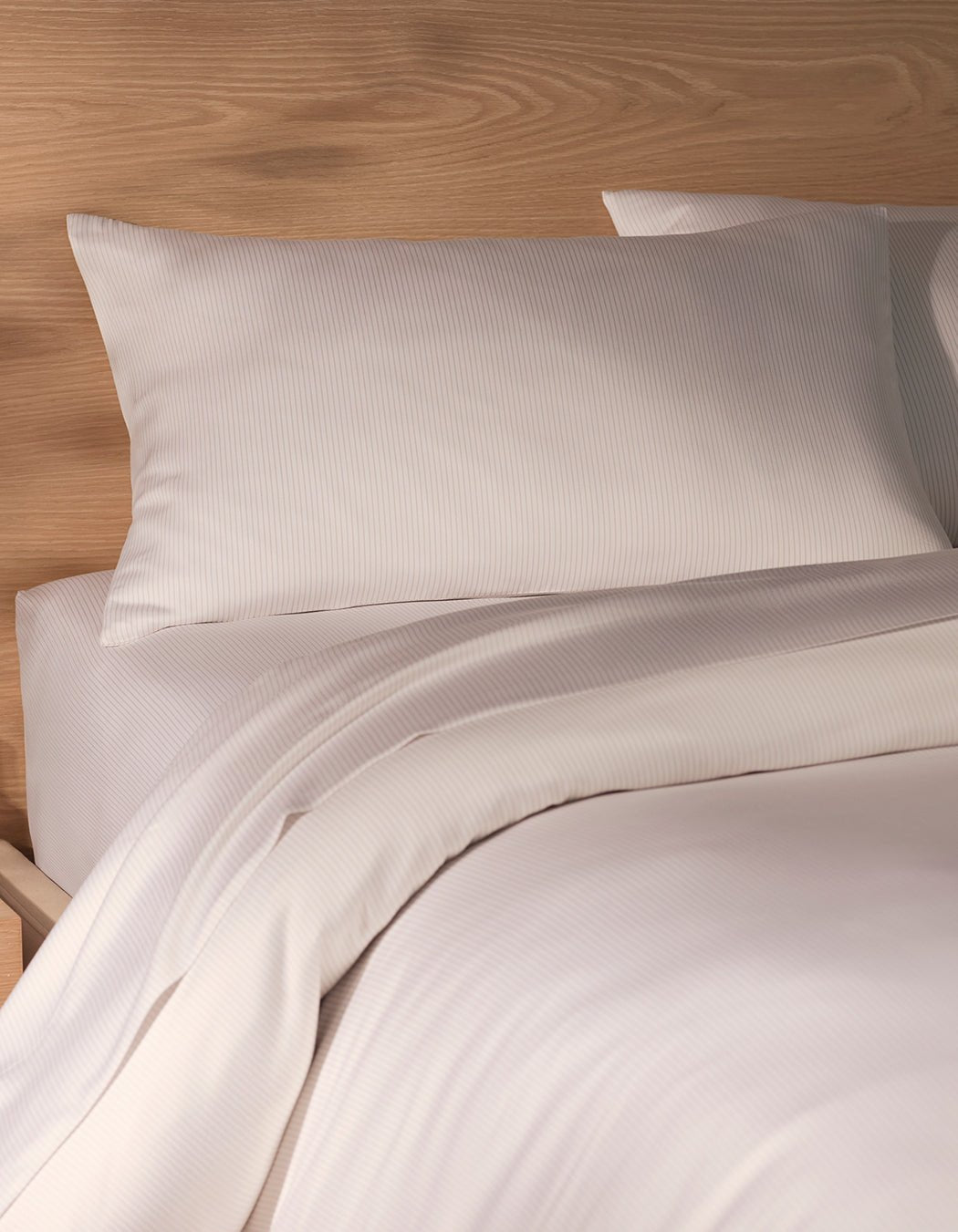 A neatly made bed features Cozy Earth Bamboo Pillowcases in white with subtle stripes, resting against a light wooden headboard. The blanket is slightly folded back, showcasing the smooth and inviting bed surface.|Color:Driftwood Pinstripe