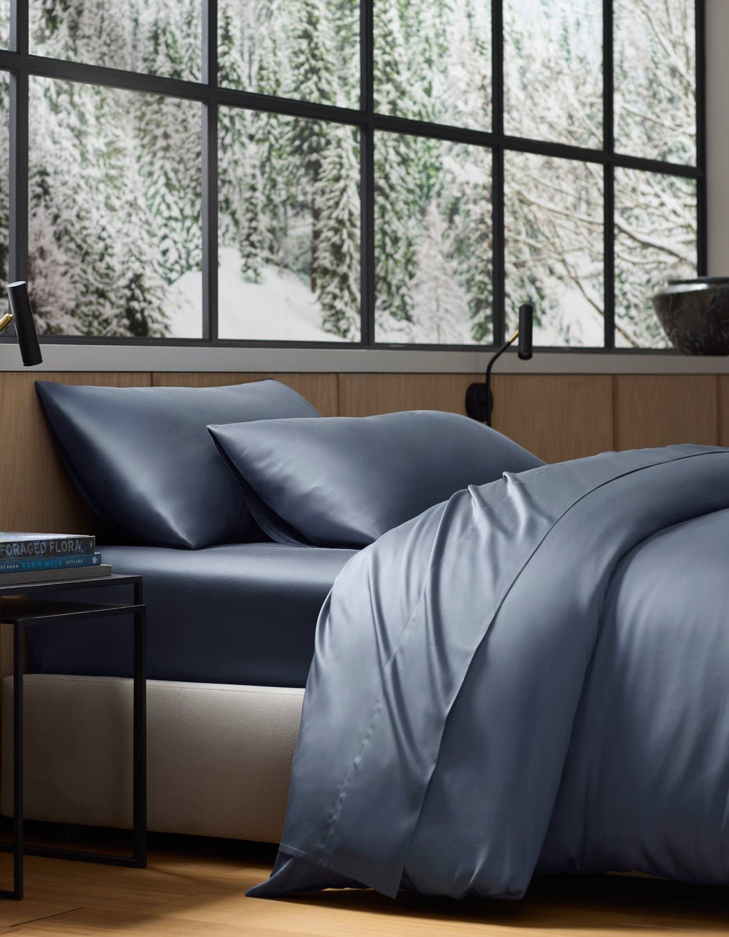 A neatly made bed featuring the Cozy Earth Bamboo Flat Sheet in dark blue sits by a large window overlooking a snowy forest. The room has wooden walls, a black lamp, and a small bedside table.