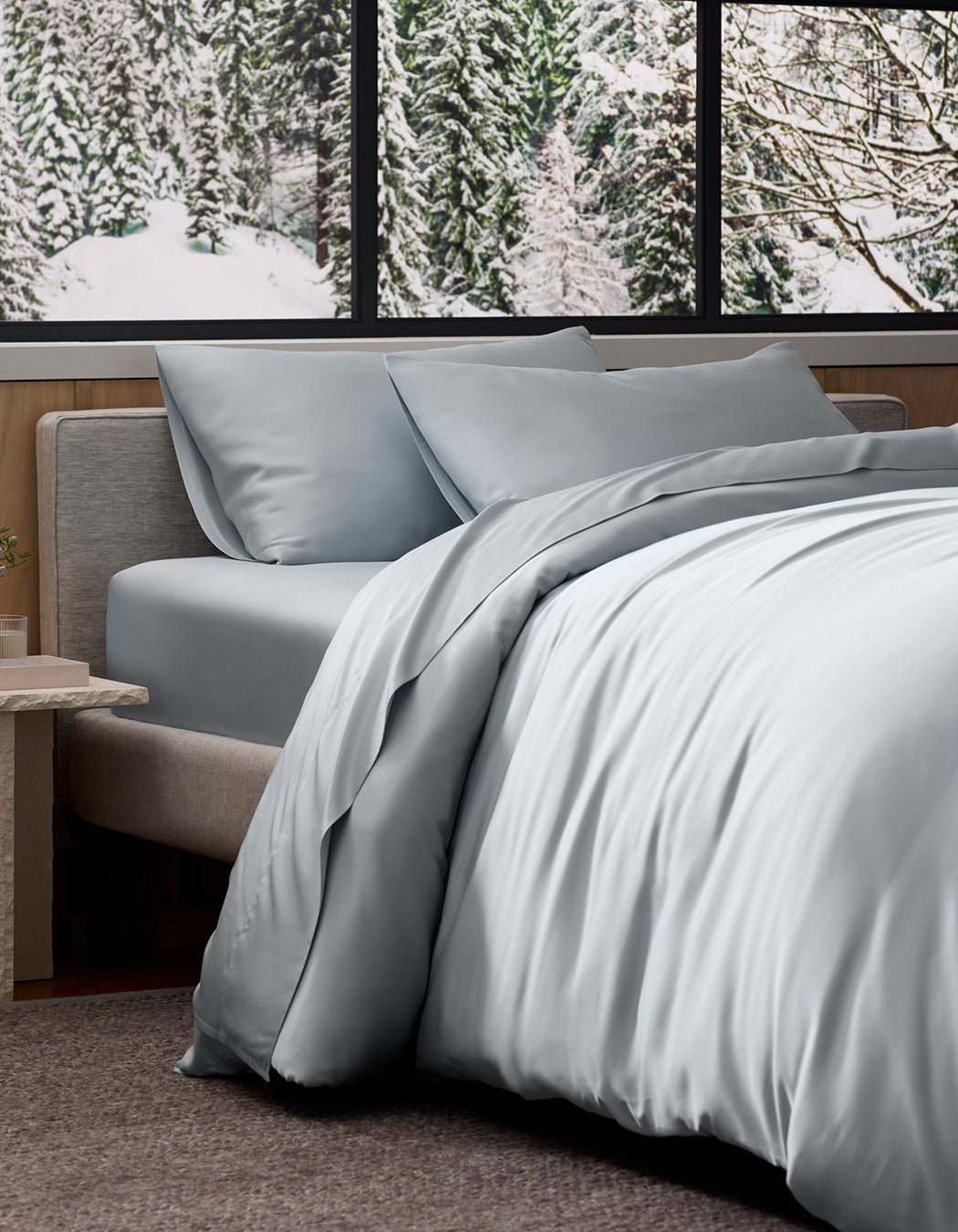 A neatly made bed featuring Cozy Earth's Bamboo Bedding Deluxe Bundle - Silk in light gray sits by a window overlooking snowy trees, creating a cozy, modern ambiance.