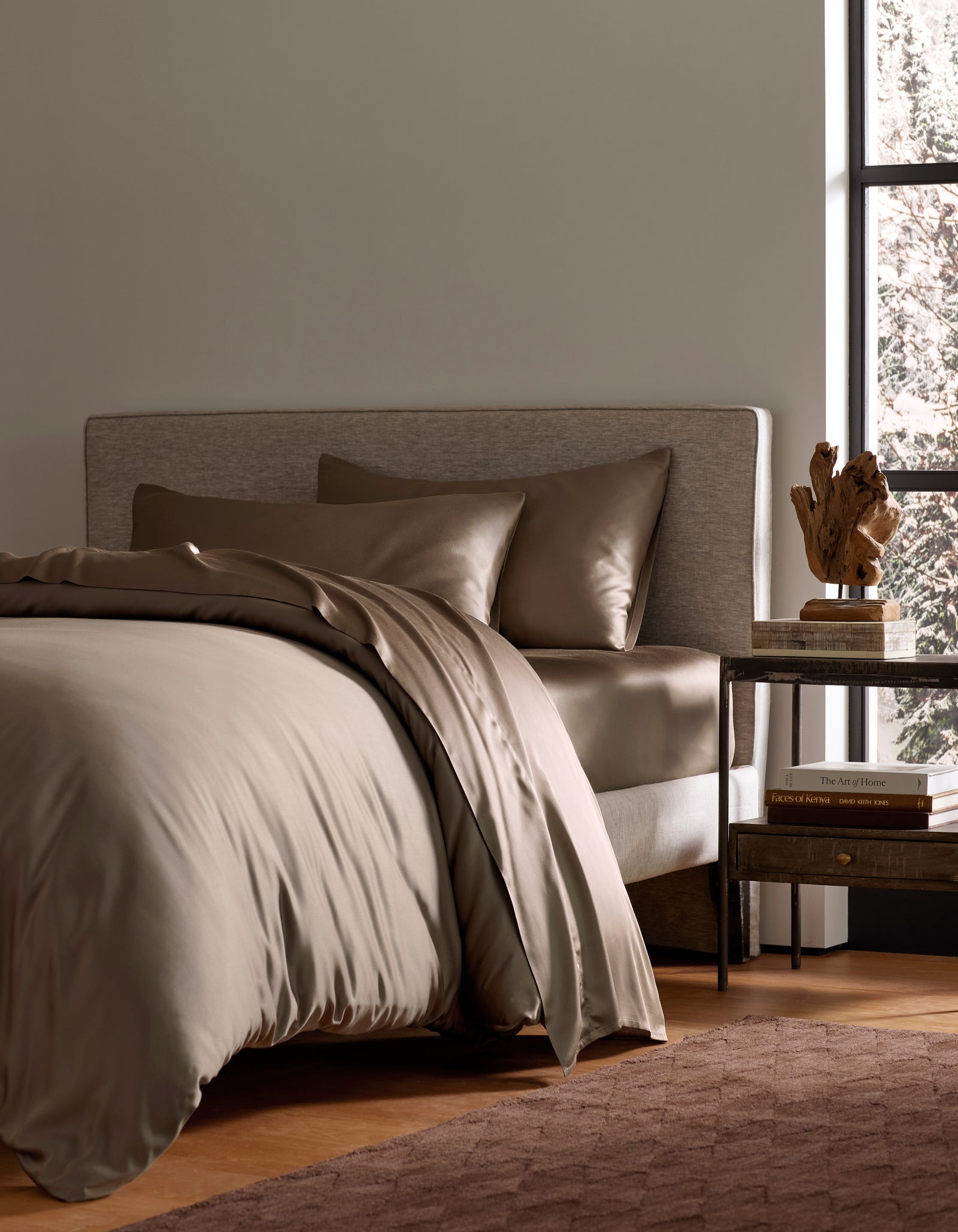 A modern bedroom features a neatly made bed with Cozy Earth’s Bamboo Bedding Deluxe Bundle in taupe, paired with pillows. A wooden nightstand nearby holds books, a lamp, and decor, as sunlight adds warmth to the inviting space.