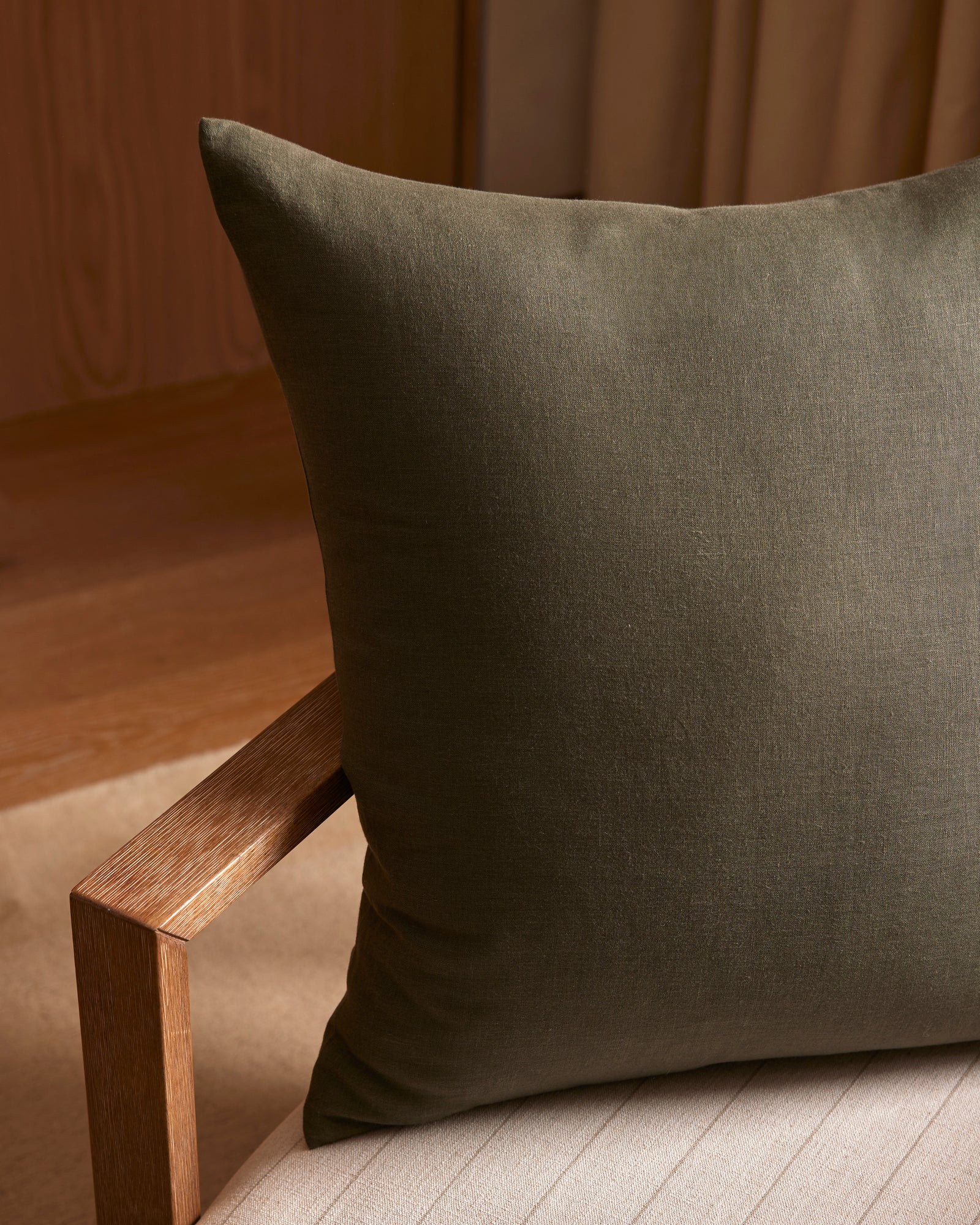 A Cozy Earth Linen Throw Pillow in dark green sits on a light beige cushioned chair with a wooden armrest, surrounded by warm lighting and wood and fabric accents in the background.