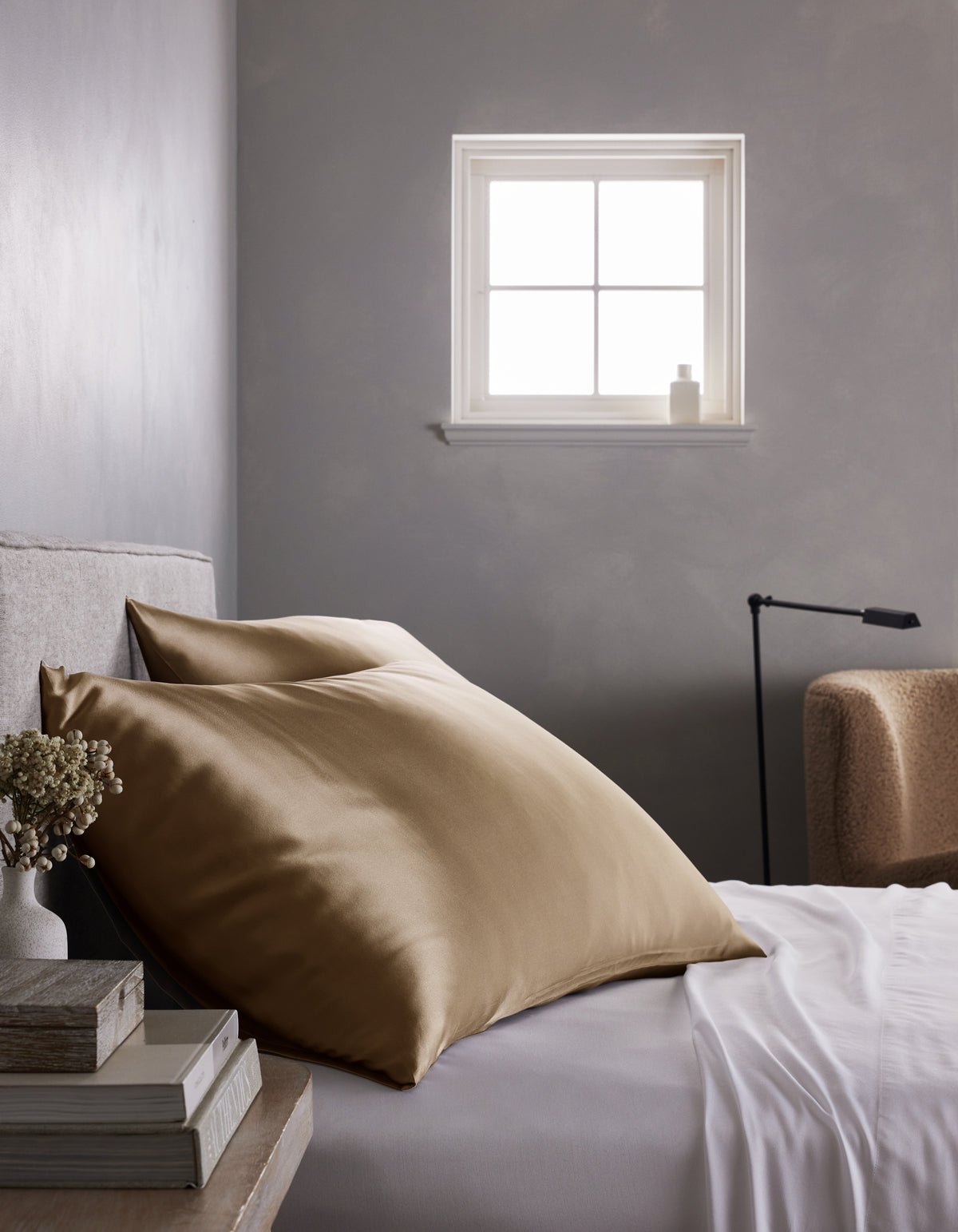 A neatly made bed features a Cozy Earth Silk Pillowcase, crisp white sheets, and a stack of books on the bedside table, all set against a gray wall with soft natural light streaming in through the window.