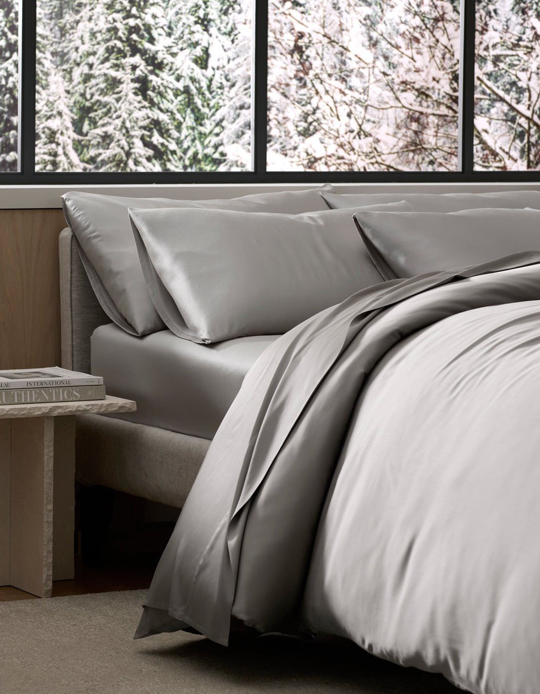 A bed dressed in Cozy Earth’s Bamboo Bedding Ultimate Bundle in light gray sits by a window with a snowy forest view, while a small bedside table holds a stack of books.