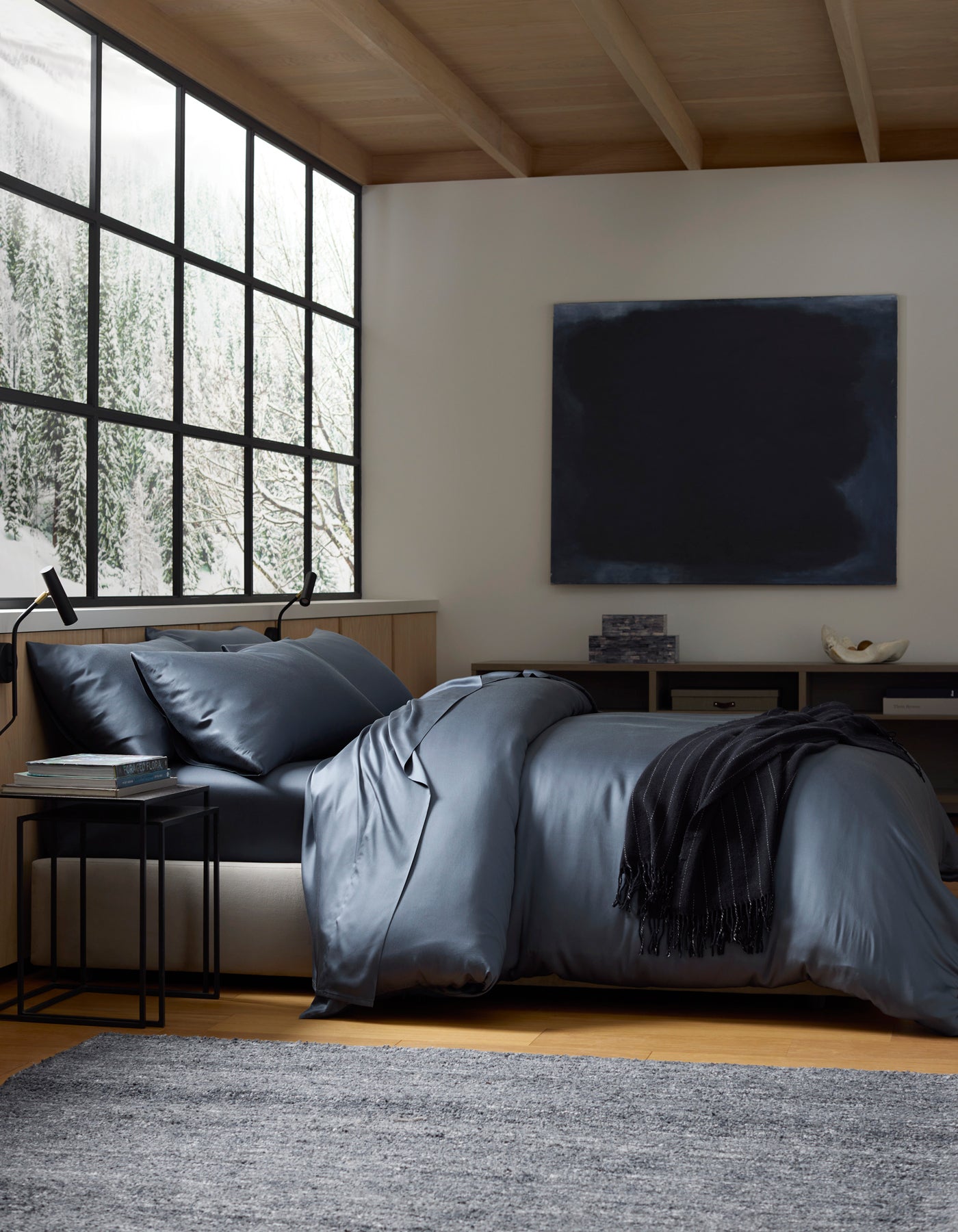 A modern bedroom with a large window showing snowy trees, a bed made with Cozy Earth's Bamboo Bedding Deluxe Bundle in dark blue, a black throw, side table, grey rug, and a dark abstract painting on the wall.|Color:Pacific Blue