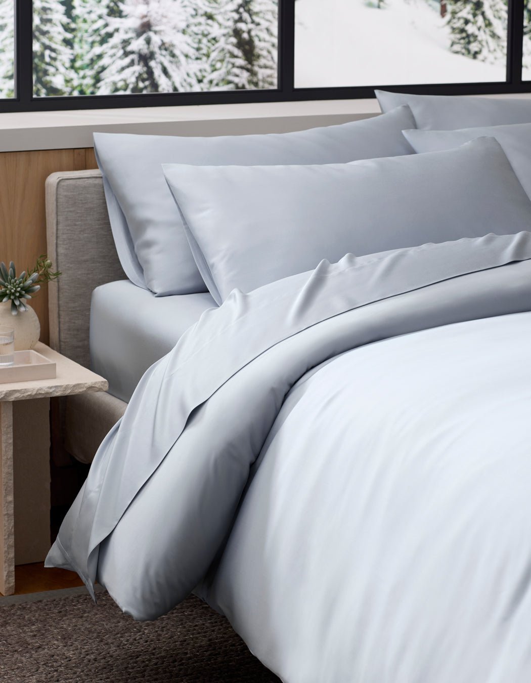A neatly made bed with Cozy Earth Bamboo Pillowcases in light gray sits beside a small table with a plant, next to a large window revealing snowy pine trees outside.