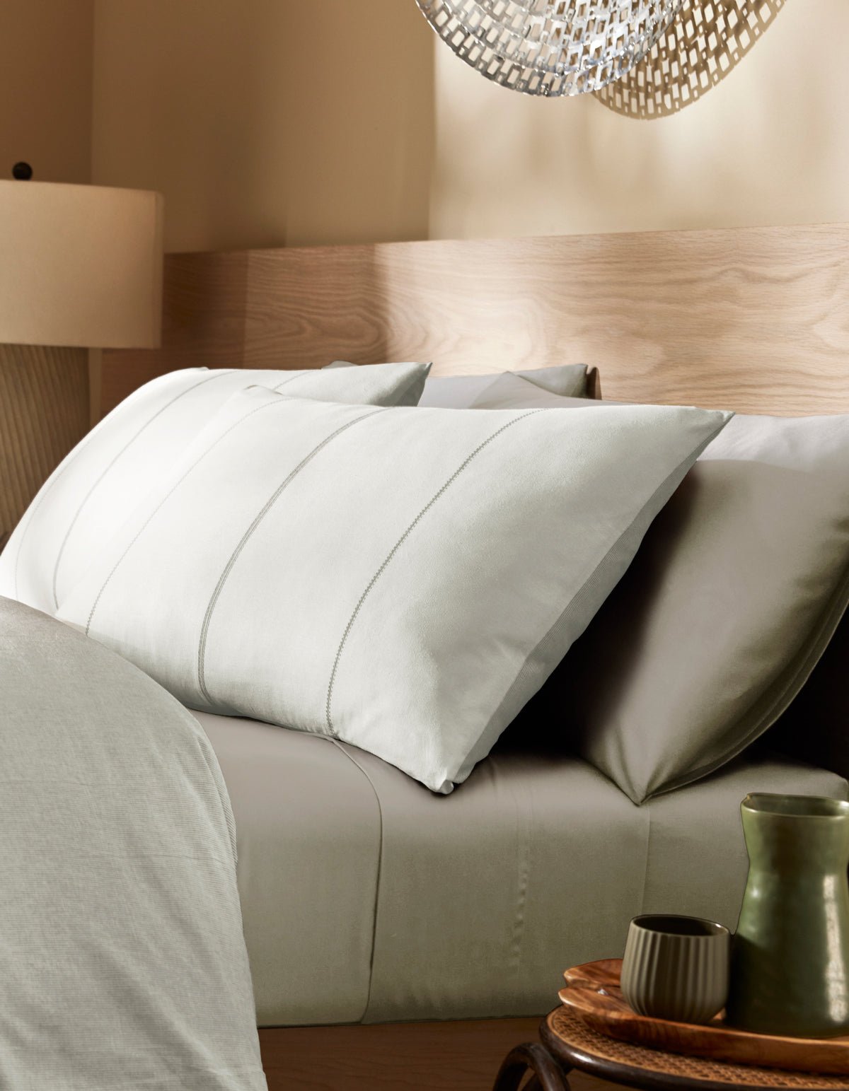 A modern bedroom featuring a neatly made bed with Cozy Earth’s Baja Double Sided Sham, light gray pillows and sheets, a wooden headboard, a bedside table holding a green cup and saucer, and a lamp under soft lighting.