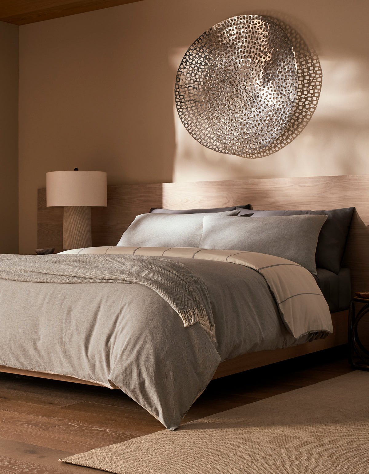 A modern bedroom featuring the Cozy Earth Baja Duvet Cover in gray, a wooden bed frame, bedside table with lamp, and metallic wall art above the bed, all creating a neutral and cozy atmosphere.