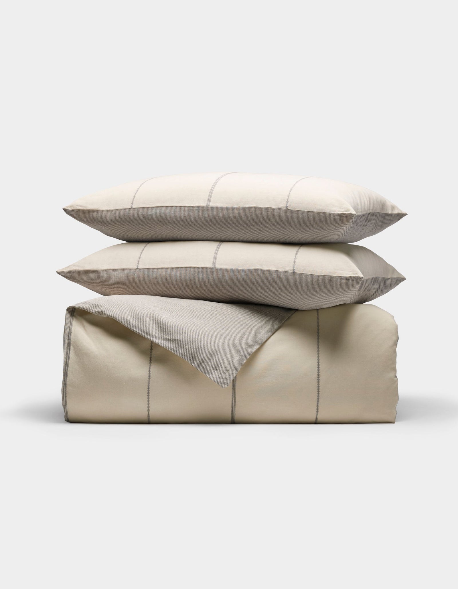 The Baja Duvet Bundle by Cozy Earth features a neatly folded beige comforter with thin light gray stripes, stacked with two matching pillows on top, all set against a plain white background.