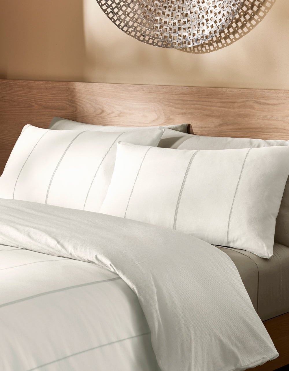 A bed featuring the Cozy Earth Baja Duvet Cover in white, with subtle stripe patterns on the bedding and pillows, is set against a light wooden headboard and beige wall adorned with metallic wall art.