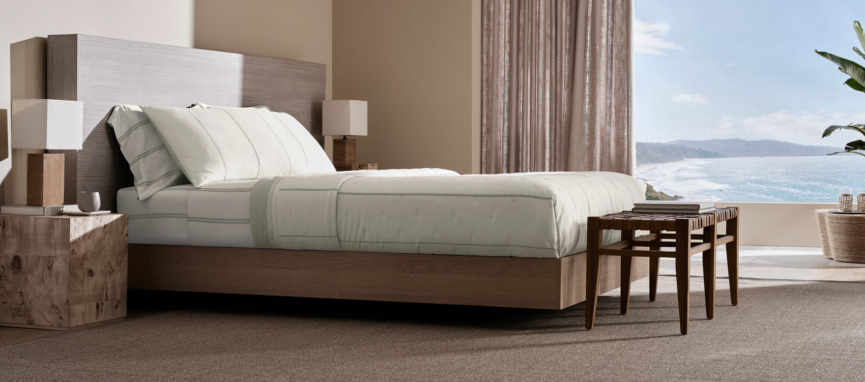 A modern bedroom featuring the Cozy Earth Baja Bedding Quilt bundle in Laurel Bone, with a large bed, wooden furniture, floor-to-ceiling curtained windows, and a scenic ocean view in the background.