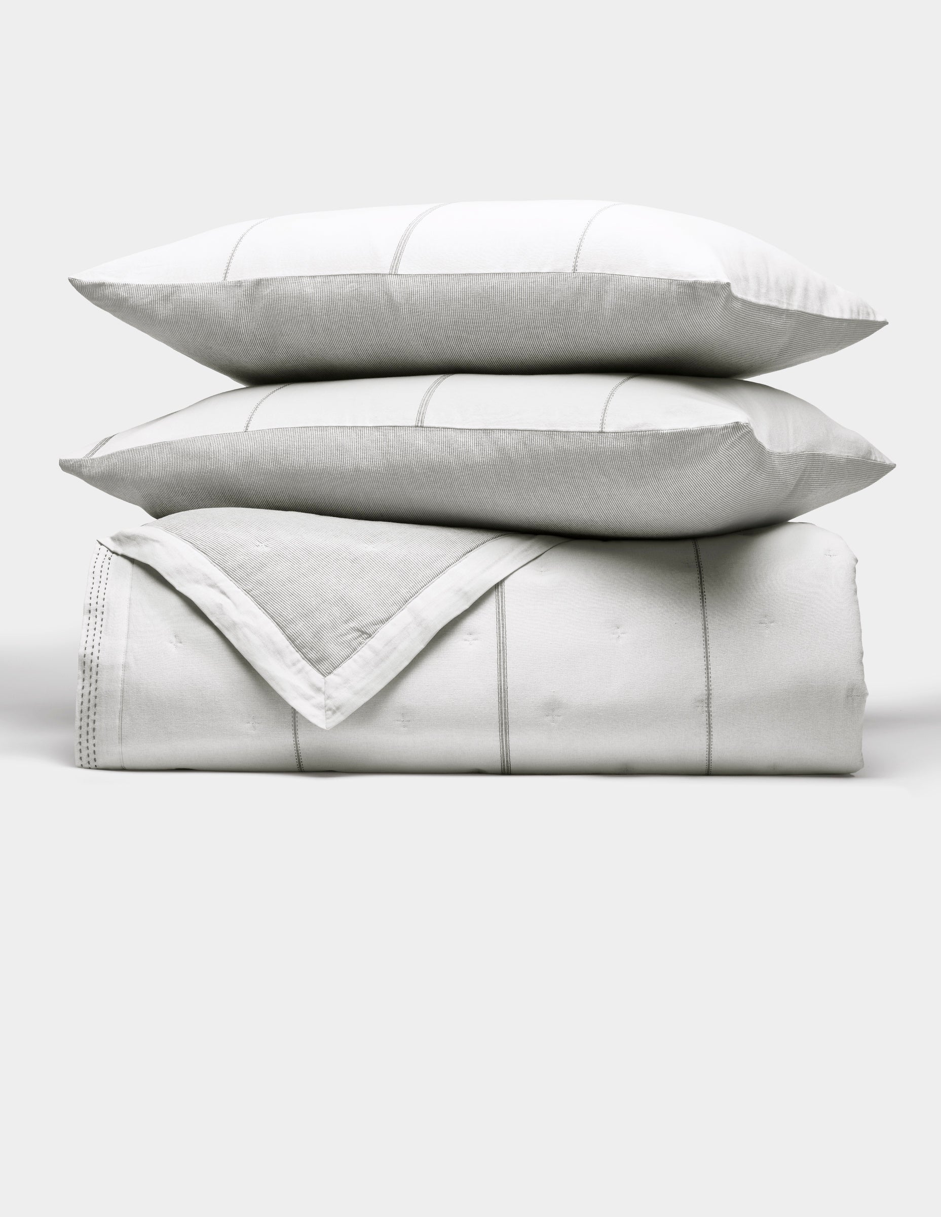 The Cozy Earth Baja Quilt Bundle features two white pillows with subtle gray stripes stacked atop a matching folded quilt, all set against a plain light background.|Color:Laurel Stripe/Striped Bone