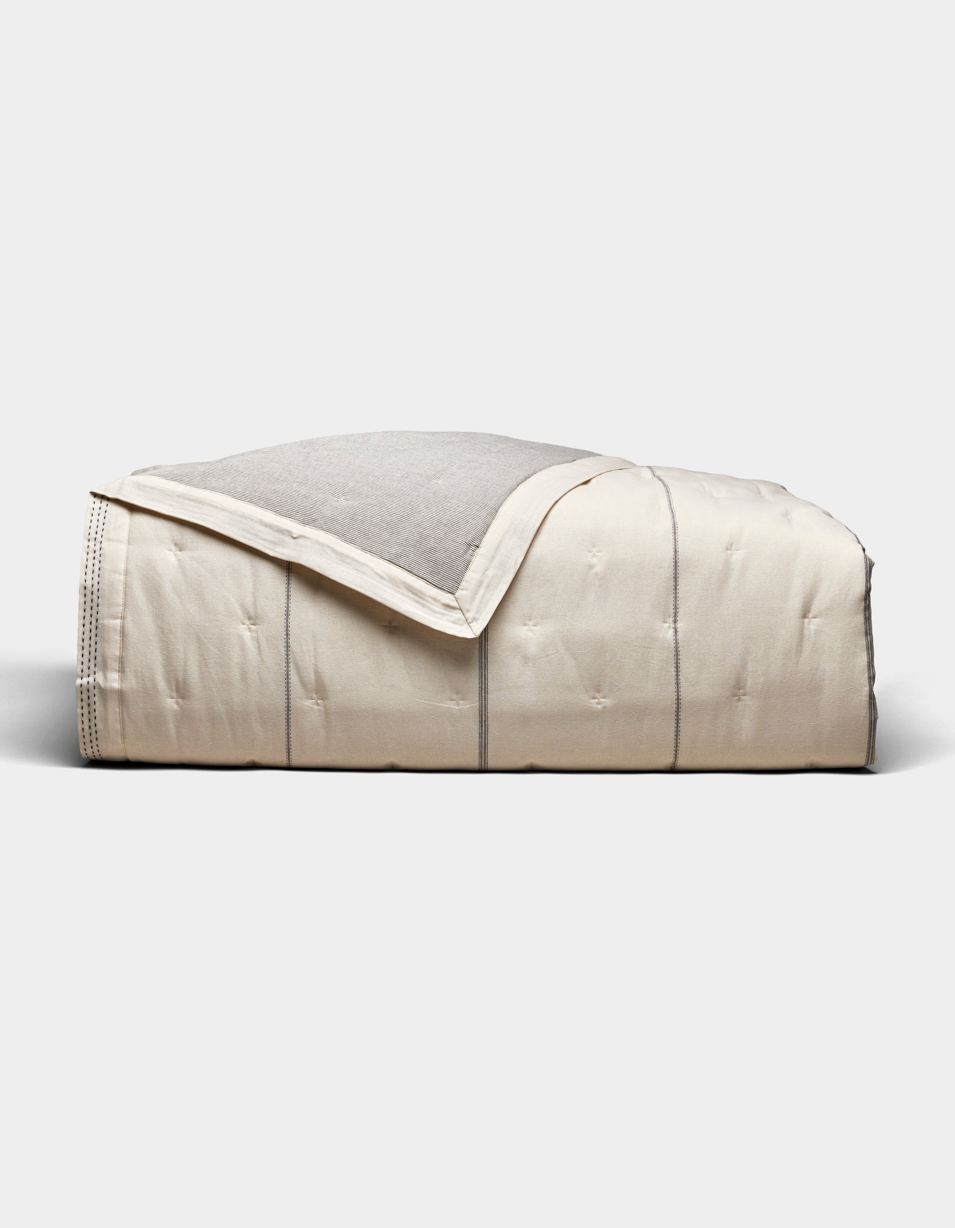 The Cozy Earth Baja Quilt, featuring a cream color with visible stitching and a light gray underside, is neatly folded and set against a plain white background.|Color:Coal Stripe/Striped Alabaster