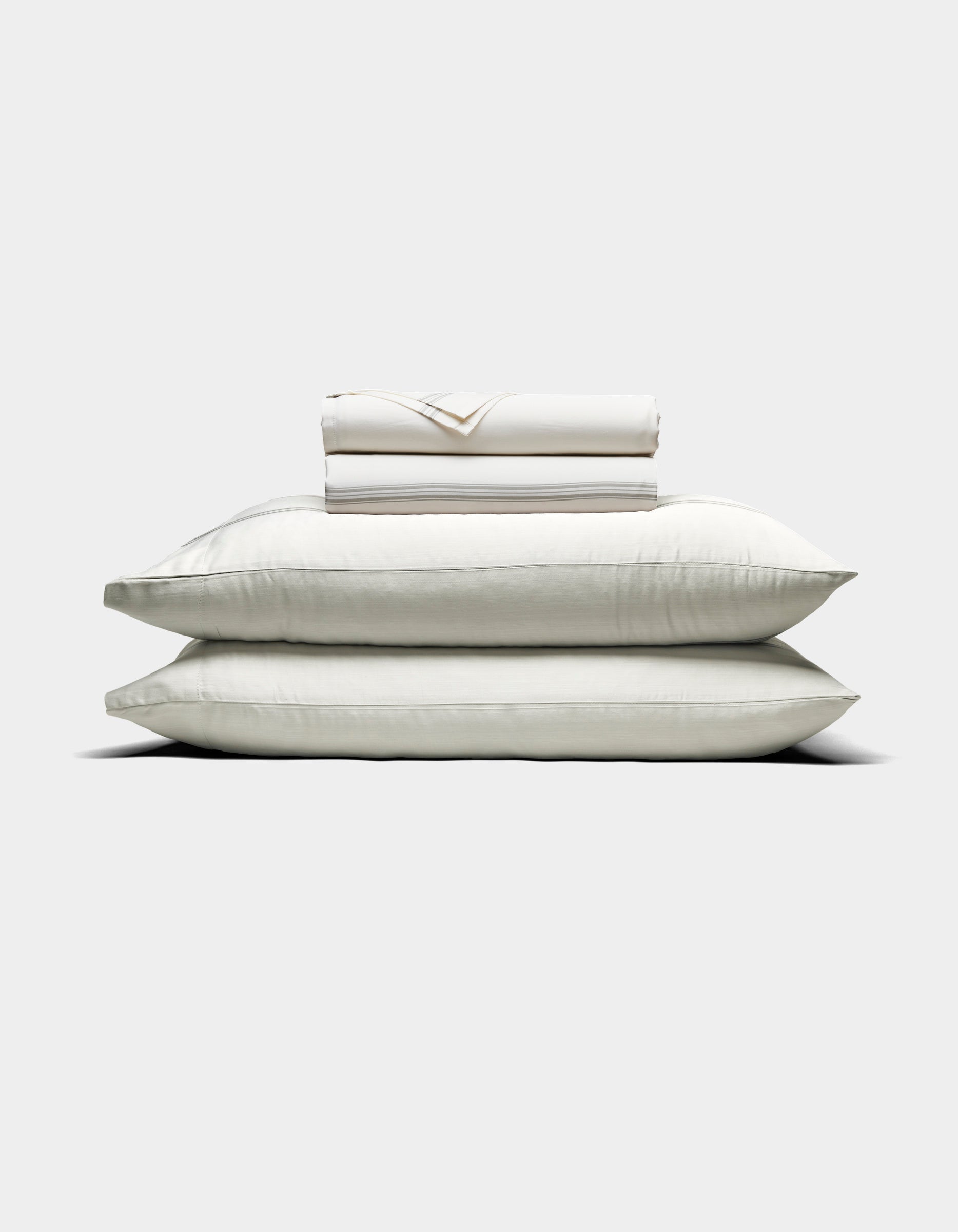 Two large white pillows are stacked with a folded Cozy Earth Bamboo Sheet Set and pillowcases placed on top, all arranged against a plain light background.|Color:Baja Stripe in Laurel