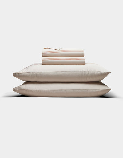 Two cream-colored pillows are stacked with Cozy Earth’s Bamboo Sheet Set, neatly folded on top, all arranged against a plain white background.|Color:Baja Stripe in Maple