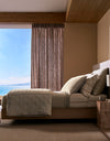 A modern bedroom showcases the Cozy Earth Baja Quilt Bundle on a neatly made bed in neutral tones, paired with a wooden nightstand and beige curtains framing an ocean view.