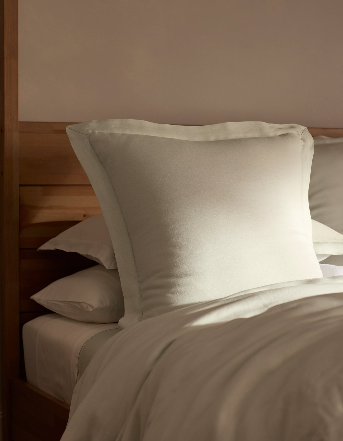 A bed featuring Cozy Earth Linen Shams is neatly made, with a large pillow propped against a wooden headboard and bathed in soft, natural light.