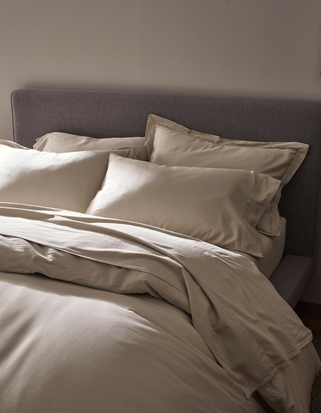 A neatly made bed features the Cozy Earth Soft-Wash Cotton Duvet Cover in cream, set against a grey upholstered headboard and softly lit for a cozy, minimalistic bedroom ambience.