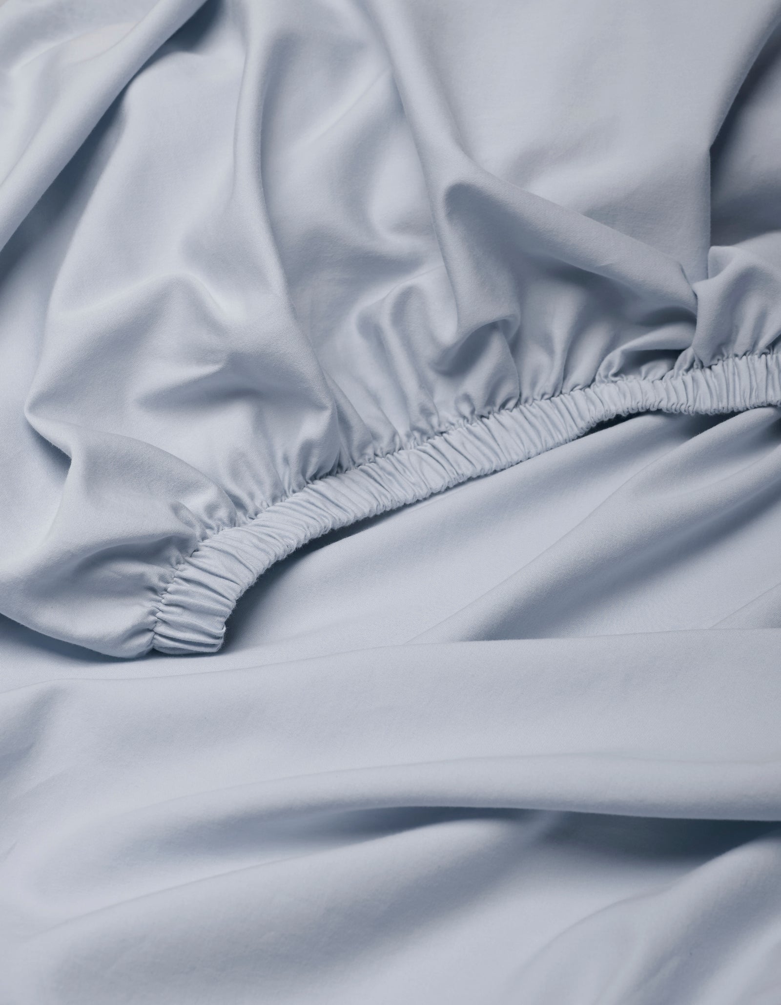The Cozy Earth Soft-Wash Cotton Fitted Sheet features a light blue color, gathered elastic edge, and close-up view highlighting its smooth texture and soft folds.